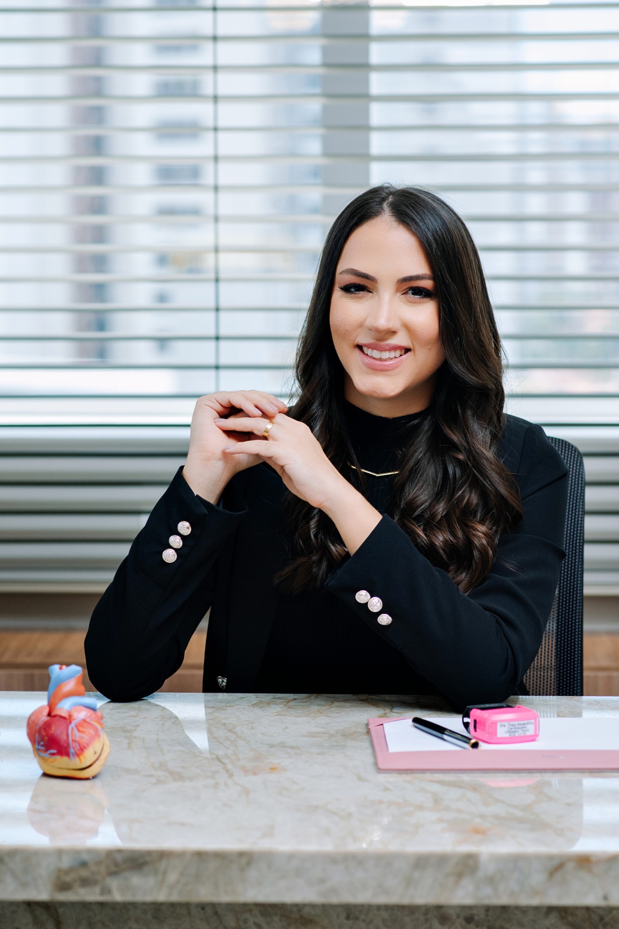 Dra. Thaís Alexandrino sentada à mesa do consultório com modelo anatômico do coração